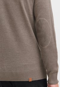 Suéter marrón claro hecho de tela suave, con puños y dobladillo acanalados, coderas y una pequeña etiqueta con logo en la parte inferior.