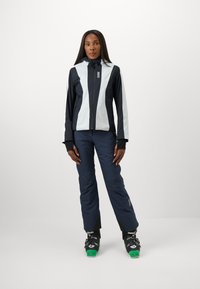 Ski jacket in navy and light blue with a zip front and hood. Paired with navy ski pants and black ski boots featuring green accents.