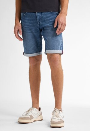 Homme portant un short en denim bleu retroussé et des baskets blanches, debout devant un fond uni.