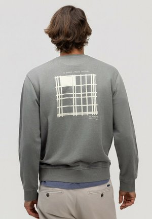 Hombre con cabello castaño que lleva una sudadera gris con patrón de cuadrícula blanca y texto en la espalda, pantalones beige, de pie contra un fondo liso.