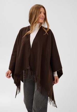 Femme portant un châle à franges marron foncé sur un col roulé blanc et un pantalon gris, regardant sur le côté et souriant sur un fond uni.
