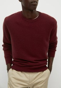 Maglione a maglia bordeaux con maniche lunghe, polsi e orlo a coste, scollatura rotonda, indossato sopra pantaloni beige. Texture morbida e design aderente.