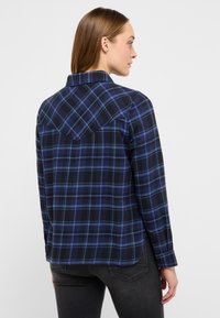 Zils un melns pārbaudīts krekls ar garām piedurknēm, apkakli un izliekta apakša. Aizmugurē ir aiziņa, un aprocēm ir pogas. Izgatavots no kokvilnas materiāla.