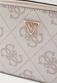 Jasnobeżowa torebka o fakturowanej powierzchni, ozdobiona subtelnym wzorem z logo, złotym trójkątnym emblematem oraz zamkiem błyskawicznym.