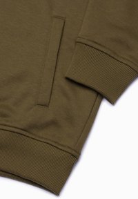 Olivenfärgad sweatshirt-tyg med en slät textur; har ribbade ärmslut och en vinklad fickdetalj framtill.