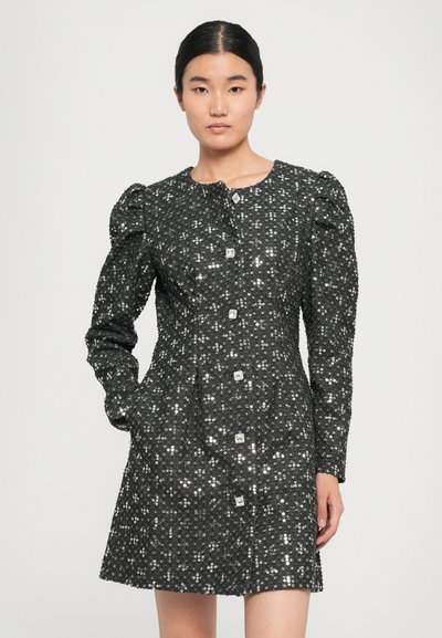 Woman wearing a long-sleeve black dress with puffed shoulders, sparkling sequins, and square decorative buttons down the front.
