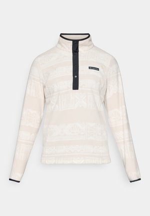 Plysjakke i lys beige med mønstrete tekstur, med høy krage, knappeåpning og marineblå detaljer.