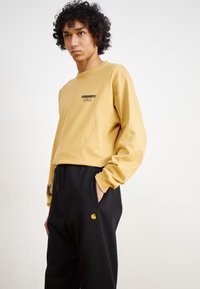Long-sleeve, mustard-yellow cotton shirt with black logo. Black pants featuring a small yellow logo and pocket embroidery with blue accents.