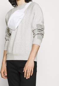 Ljusgrå sweatshirt med ribbad mudd vid ärmslut och nederkant, med en framficka. En vit axelväska bärs över bröstet. Svarta byxor.