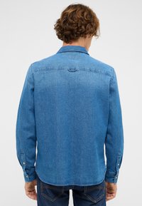 Hele sinise teksariidest särk, millel on nööbitavad mansetid, pikad varrukad ja klassikaline krae. Omadused hõlmavad sirget seljaosa ja nähtamatuid mustreid.