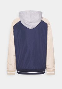 Veste bleu marine avec manches beiges, capuche gris clair, poignets côtelés et ourlet rayé bleu marine et blanc vue de dos.