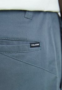 Pantalon gris Volcom en tissu texturé, avec une poche arrière ornée d'une couture diagonale et une petite étiquette de la marque.