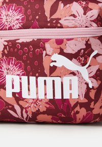 Marooni ja roosa lillemustriga kangast kott, millel on silmatorkav valge "PUMA" logo ja logode siluett tekstuurse tausta ees.