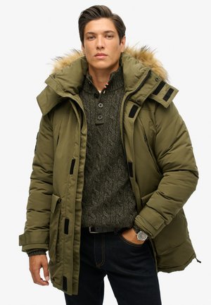 Chaqueta acolchada verde oliva con capucha forrada de piel, múltiples bolsillos y detalles en negro. Llevada sobre un suéter oscuro de gris texturizado.