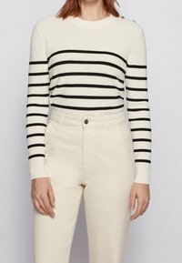 Pull rayé crème avec des rayures noires, en maille, col rond, et détail de boutons sur l'épaule. Associé à un pantalon de couleur claire.