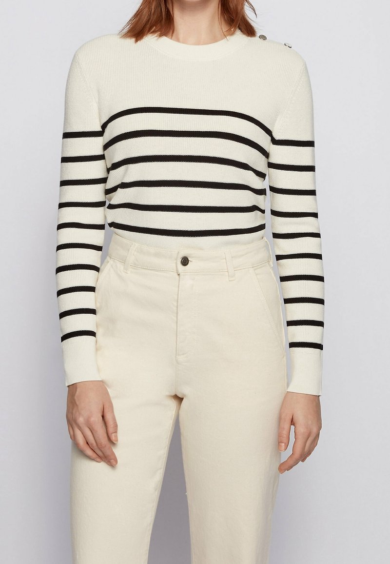 Pull rayé crème avec des rayures noires, en maille, col rond, et détail de boutons sur l'épaule. Associé à un pantalon de couleur claire.