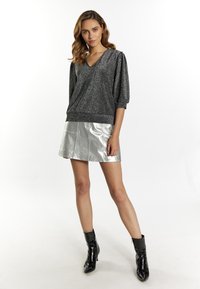 Glänzender schwarzer Kurzarm-Pullover mit V-Ausschnitt. Kombiniert mit einem metallisch-silbernen Minirock und schwarzen Lack-Ankle-Boots. Glatte Textur.
