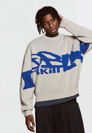 Jeune homme aux cheveux tressés portant un pull crème avec un motif de texte abstrait bleu, pantalon noir, posant avec une main sur la tête.