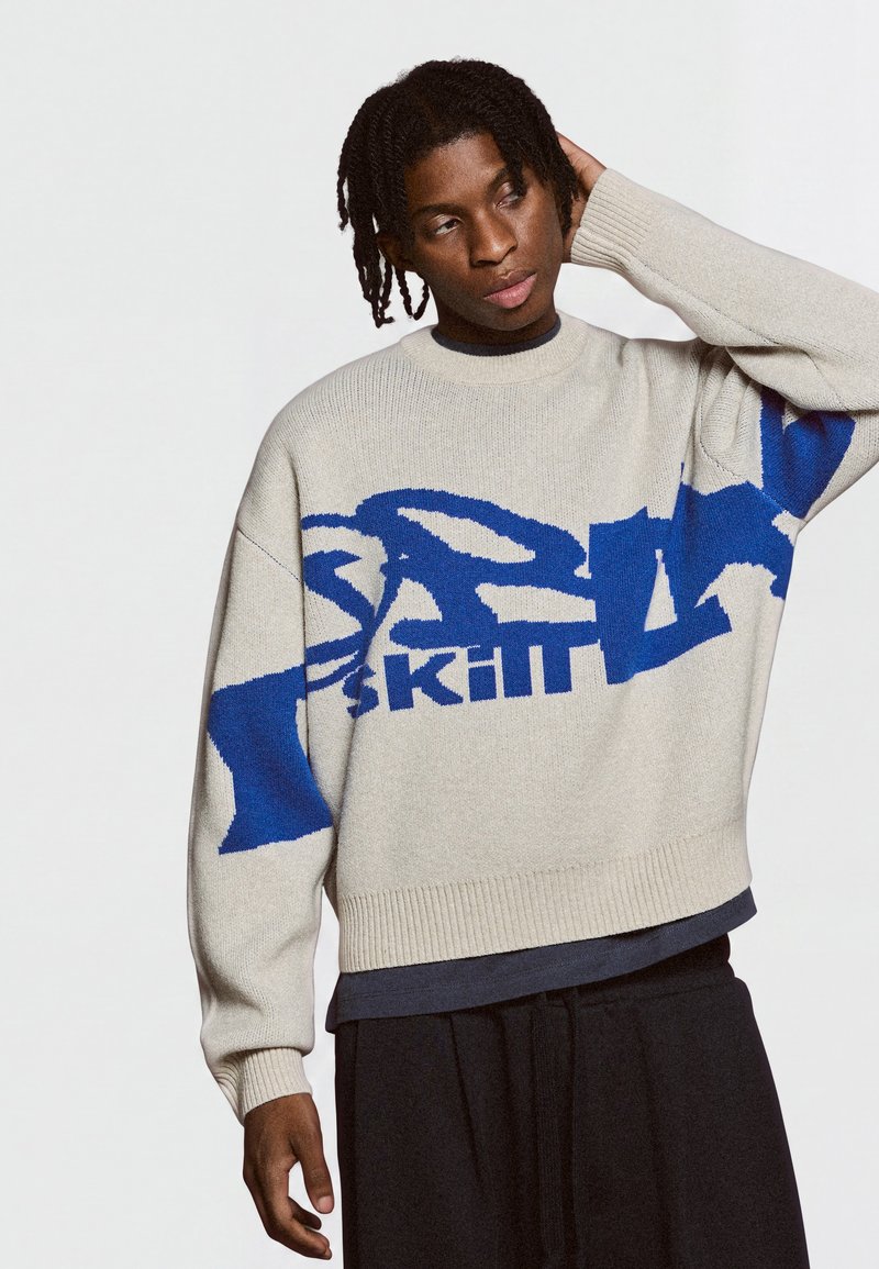 Jeune homme aux cheveux tressés portant un pull crème avec un motif de texte abstrait bleu, pantalon noir, posant avec une main sur la tête.