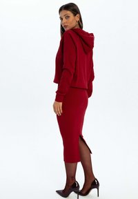 Czerwona krótka bluza z kapturem i sznurkiem, w zestawieniu z dopasowaną spódnicą midi z bocznym rozcięciem. Noszona z czarnymi wysokimi obcasami. Gładka tekstura.