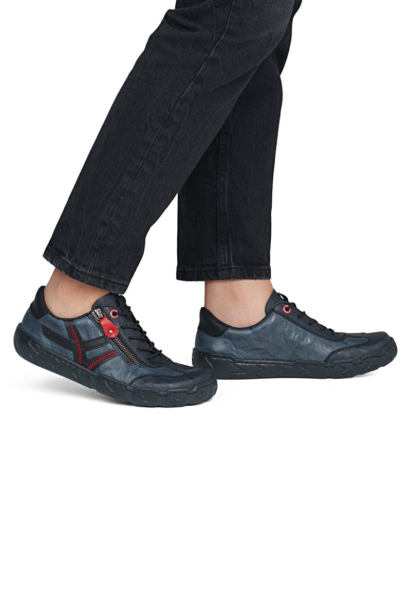 Pieds portant des chaussures décontractées en cuir bleu foncé avec fermeture éclair rouge et détails en lacets, associés à un pantalon noir raccourci sur fond blanc.