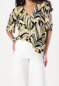 Blusa a maniche corte con scollo a V, realizzata in tessuto leggero con un motivo di foglie multicolore nei colori verde, nero, beige e marrone.