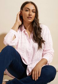 Camicia button-up rosa chiaro con ricami a occhio, maniche lunghe e vestibilità rilassata. Indossata con pantaloni in denim scuri.