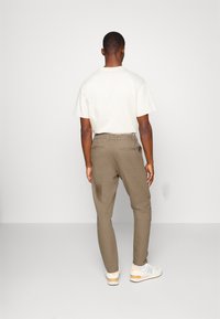 Homme de dos portant un t-shirt blanc, un pantalon ajusté olive, et des baskets blanches avec des accents beiges sur un fond gris.