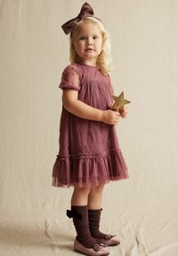 Lila Kleid mit besticktem Blumenmuster und Tüllbesatz. Das Kind trägt ein passendes Stirnband, kniehohe Socken mit Schleifen und flache Schuhe.