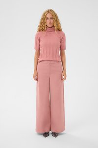 Kvinde med krøllet blond hår iført en lyserød ribbet turtleneck kortærmet top og brede lyserøde bukser, stående mod en hvid baggrund.