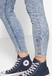 Light blue, slim-fit jeans with a faded texture and button accents at the ankle, paired with dark blue canvas high-top sneakers.