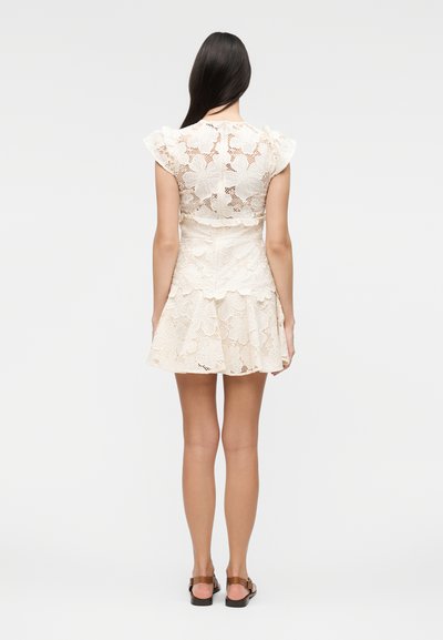 Γυναίκα γυρισμένη με την πλάτη, φορεί κοντό, κρεμ δαντελένιο φόρεμα με λουλουδένια σχέδια και κοντά φουσκωτά μανίκια, συνδυασμένο με καφέ σανδάλια.