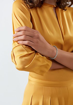Gelbe Satinbluse mit Puffärmeln und einem Schlüsselloch-Ausschnitt. Ein silbernes geflochtenes Armband schmückt eine Hand, die entspannt auf dem Arm liegt.