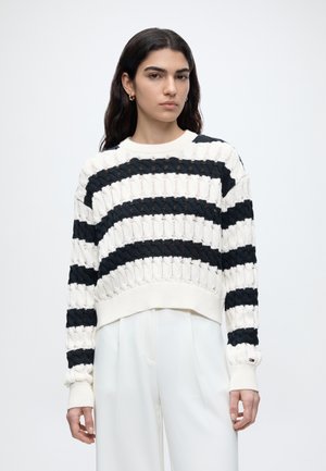 Kobieta o długich ciemnych włosach, ubrana w biało-czarny prążkowany sweter i białe szerokie spodnie z wysokim stanem, stoi na jednolitym tle.