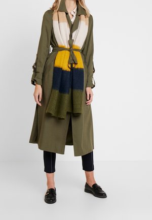 Femme portant un trench-coat ceinturé vert olive, une écharpe rayée multicolore, un pantalon noir court et des mocassins noirs sur fond blanc.