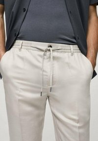 Lichtbeige joggings met een strikband in de taille, zijzakken en een gladde textuur, gedragen met een grijze top en een donker overhemd met knopen.