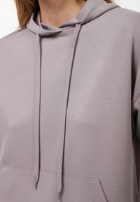 Jasnofioletowy hoodie z kangurzą kieszenią, sznurkami i miękką teksturą. Design jest prosty, bez widocznych wzorów czy logo.