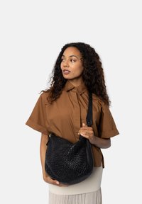Vrouw met krullend haar in een bruine blouse en lichte rok houdt een zwart geweven leren schoudertas vast, terwijl ze naar links kijkt.