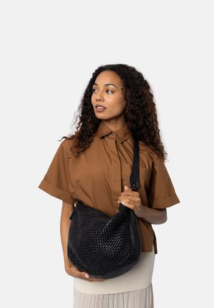 Donna con capelli ricci, che indossa una camicia marrone e una gonna chiara, tiene una borsa a tracolla in pelle intrecciata nera, guardando verso sinistra.