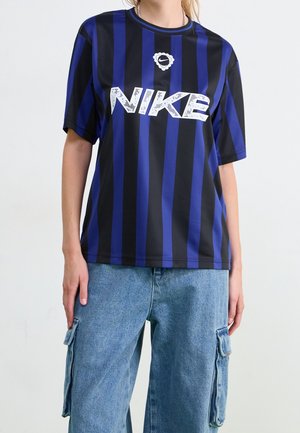 Personne portant un t-shirt Nike à rayures verticales noires et bleues et un pantalon cargo en denim bleu clair avec des poches latérales, sur un fond uni.