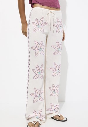 Pantalones blancos holgados con bordado floral púrpura y costuras laterales, combinados con sandalias blancas y una blusa corta púrpura.