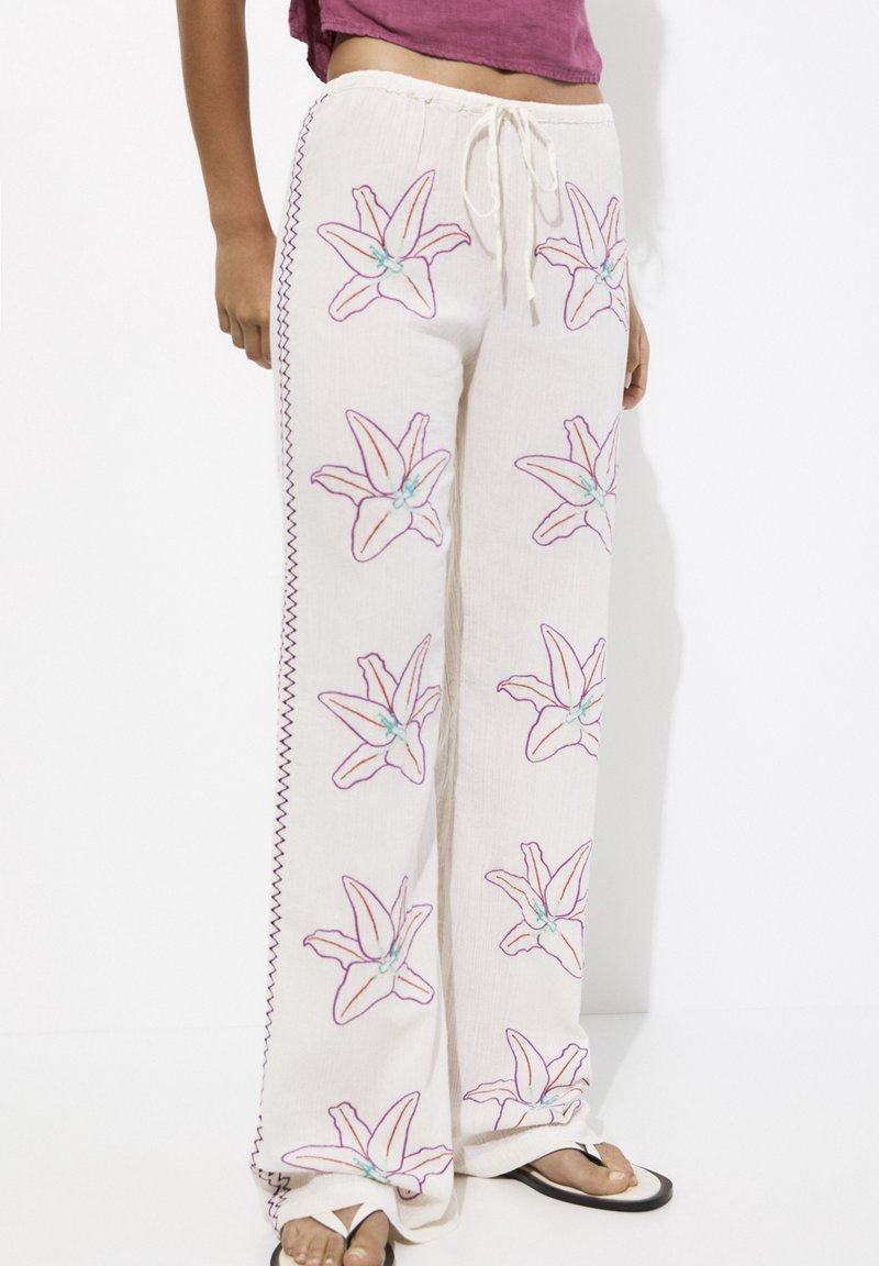 Pantalon blanc ample avec broderie florale violette et surpiqûres latérales, porté avec des sandales blanches et un haut court violet.