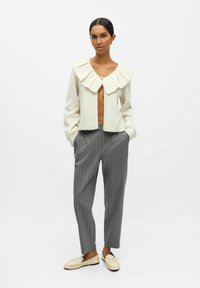 Cremfarbener Cardigan mit übergroßem Rüschensaum, offener Vorderseite und Knopfverschluss, kombiniert mit grauen Nadelstreifenhosen und hellen Loafers.