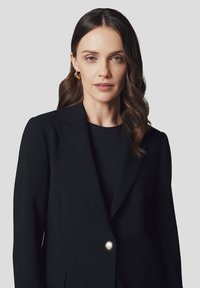 Czarna, dopasowana marynarka z pojedynczym srebrnym guzikiem, z klapami i elegancką sylwetką; gładki materiał i wyrazisty krój.