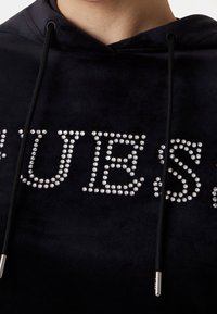 Sweat à capuche en velours noir avec lettrage en strass. Comprend des cordons noirs avec embouts métalliques. Texture douce et ornements brillants.