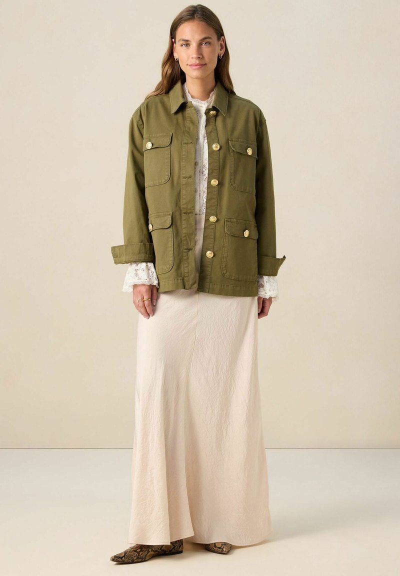 Veste utilitaire vert olive avec quatre poches et boutons dorés, superposée à une longue jupe légère et texturée. Bottines à enfiler brunes à motif.