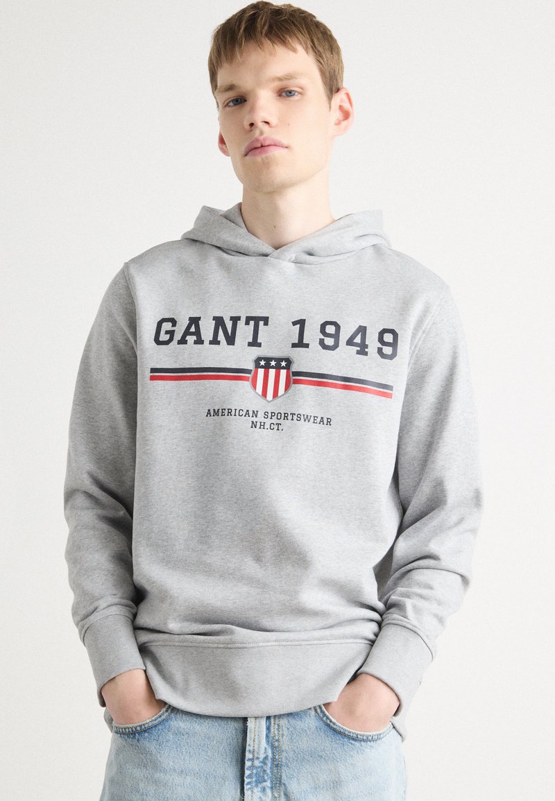 Hoodie gris en tissu doux avec le logo "GANT 1949", un graphique en forme de blason, et des rayures rouges et bleu marine, associé à un jean bleu clair.