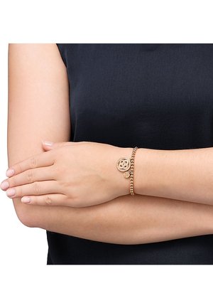 Personne portant un haut noir sans manches, les bras croisés, avec un bracelet en perles dorées orné de deux breloques rondes au poignet droit.