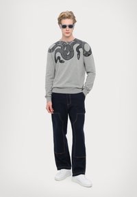 Maglione grigio con design di serpente nero sulle spalle, abbinato a pantaloni blu navy con tasche a toppa e sneakers bianche.