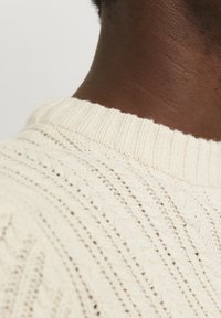 Cream-colored knitted sweater with a ribbed round collar and a textured pattern, showcasing a soft, slightly chunky fabric.